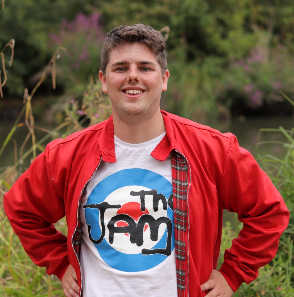 Director, Will Hextall, standing confidently and smiling with his hands on his hips, wearing a red Harrington jacket and a T Shirt, with the logo of a band called "The Jam". He stands in front of a lake with lavender distant in the background.